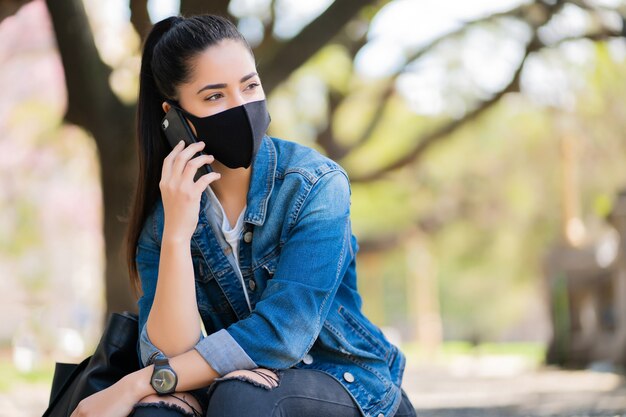 Portret młodej kobiety noszenie maski i rozmawia przez telefon, siedząc na schodach na zewnątrz. Koncepcja miejska