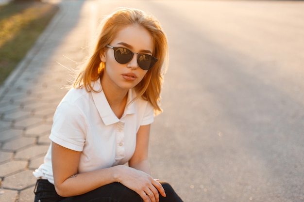 Portret młodej kobiety piękne hipster w stylowe okulary w białej koszulce polo w czarne dżinsy o zachodzie słońca. Amerykańska dziewczyna siedzi na asfaltowej drodze.