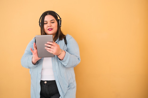 Portret młodej kobiety plus size słuchanie muzyki w słuchawkach i cyfrowy tablet na zewnątrz na żółto.