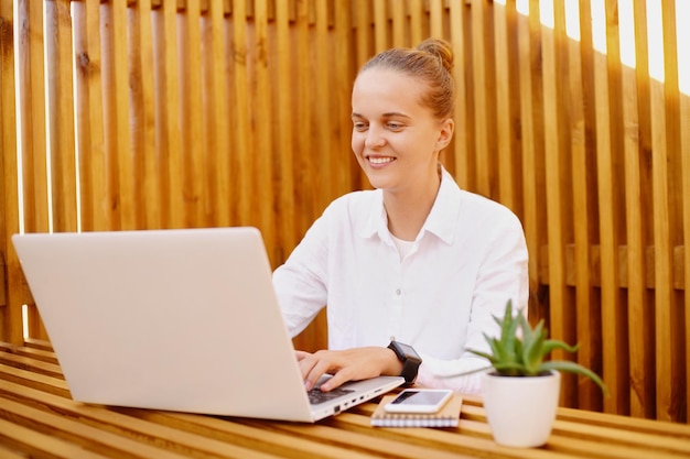 Portret młodej kobiety rasy kaukaskiej w kawiarni lub biurze, pracując na komputerze przenośnym, wyrażając pozytywne emocje podczas pisania na notebooku freelancer z fryzurą kok na sobie białą koszulkę
