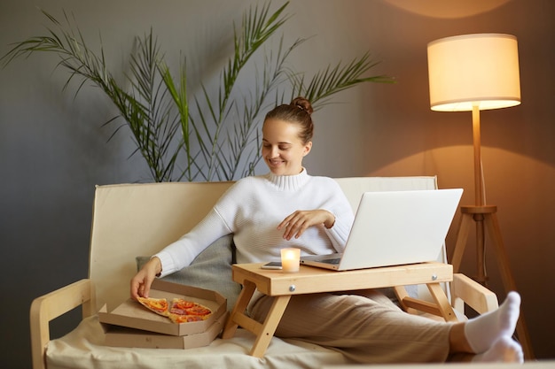 Portret młodej kobiety siedzącej na kanapie, relaksującej się podczas przeglądania Internetu w czasie wolnym w domu i jedzącej świeżo upieczoną pizzę, ciesząc się wolnym czasem w weekend
