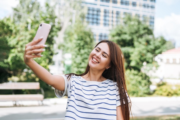 Portret młodej kobiety studentki z długimi włosami biorąc selfie z telefonem komórkowym w parku miejskim