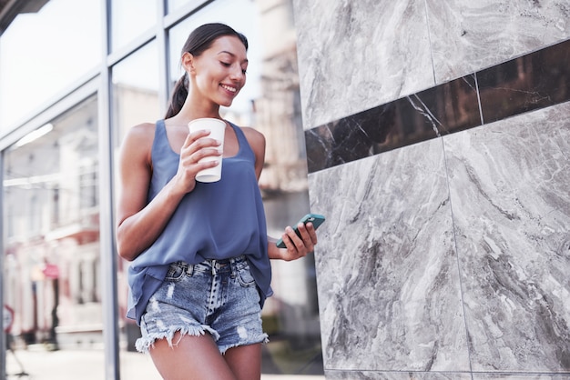 Portret młodej kobiety stylowe hipster spaceru na ulicy, ubrany w ładny modny strój, picie gorącej kawy latte i uśmiechnięty