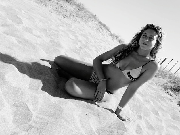 Zdjęcie portret młodej kobiety w bikini siedzącej na piasku na plaży przy jasnym niebie
