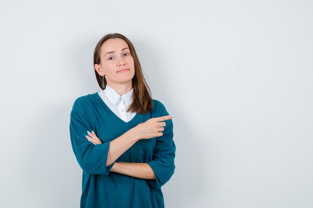 Portret Młodej Kobiety, Wskazując W Prawo W Swetrze Nad Białą Koszulą I Patrząc Pewny Siebie Widok Z Przodu