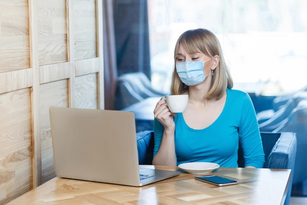 Portret młodej kobiety z chirurgiczną maską medyczną w niebieskiej koszulce siedzi, pracuje i patrzy na ekran laptopa i pije kawę. Praca na odległość w pomieszczeniach, koncepcja medycyny i opieki zdrowotnej.