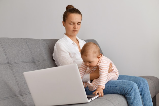 Portret młodej kobiety z laptopem siedzącej na kanapie z dzieckiem, patrzącej na ekran laptopa ze smutnym wyrazem twarzy, ubrana w białą koszulę i dżinsy, zdenerwowana i odczuwająca smutek.