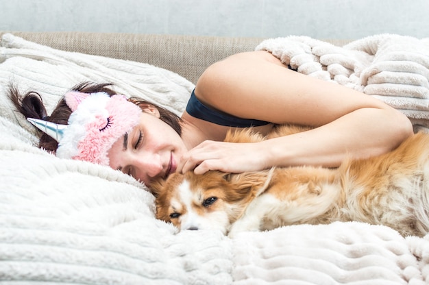 Portret młodej kobiety z maską do spania i piżamą przytulającą psa. Pojęcie odpoczynku i snu.