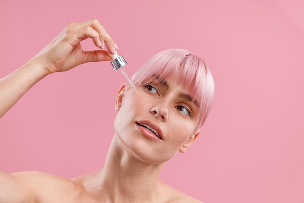 Portret młodej kobiety z różowymi włosami, która nakłada serum lub kwas hialuronowy na skórę twarzy za pomocą