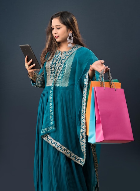 Portret młodej kobiety z telefonem komórkowym lub tabletem i torbami na zakupy w dłoniach na szarym tle