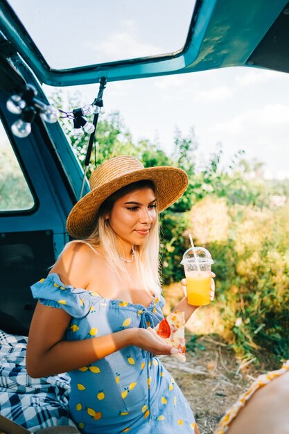 Portret młodej ładnej kobiety siedzącej na kocu w vanie i picia lemoniady i jedzenia pizzy na świeżym powietrzu w przyrodzie. Ciesz się latem, koncepcja podróży