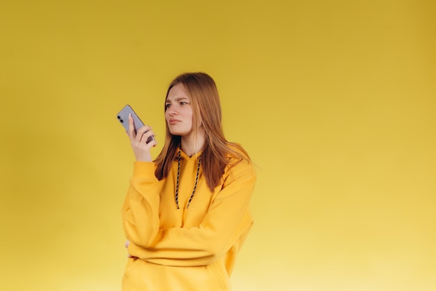 Portret młodej nastolatki trzymającej telefon dziewczyna rozmawiająca przez telefon ubrana w niezobowiązującą żółtą bluzę z kapturem w jednym tonie z tłem