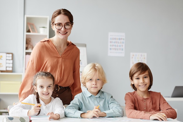 Zdjęcie portret młodej nauczycielki w okularach uśmiechającej się do kamery z grupą dzieci siedzących przy stole...