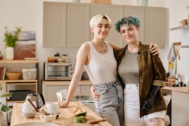 Portret młodej pary lesbijek obejmującej kamerę i patrzącej na nią podczas przygotowywania jedzenia w kuchni