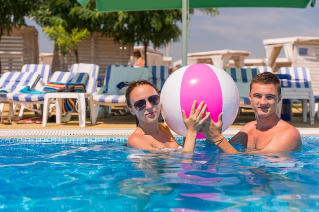 Portret Młodej Pary Na Wakacjach W Basenie, Trzymając Razem Nadmuchiwaną Piłkę Plażową W Słoneczny Dzień W Ośrodku