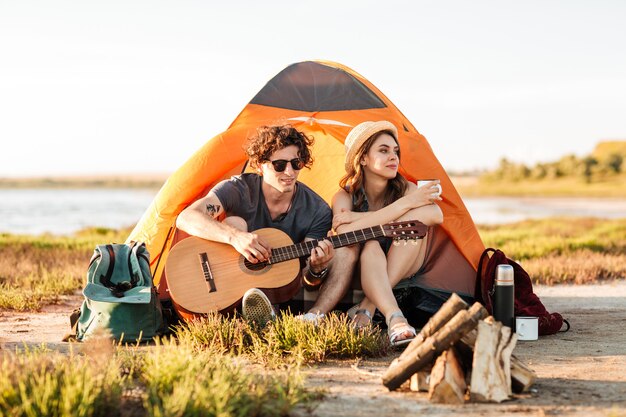 Portret młodej pary siedzi z gitarą w pobliżu ogniska podczas biwakowania