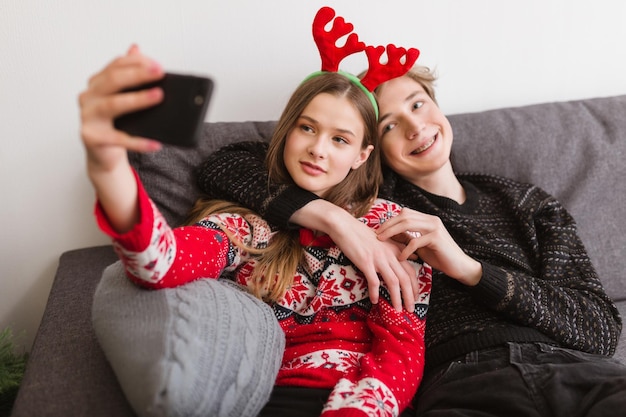 Portret młodej pary uśmiechniętej siedzącej na kanapie w domu i biorącej śmieszne selfie podczas wspólnego spędzania czasu