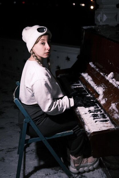 Portret młodej pianistki w białych zimowych ubraniach i czarnych rękawiczkach na świeżym powietrzu w zimie z bliska ze śniegiem na klawiszach fortepianu