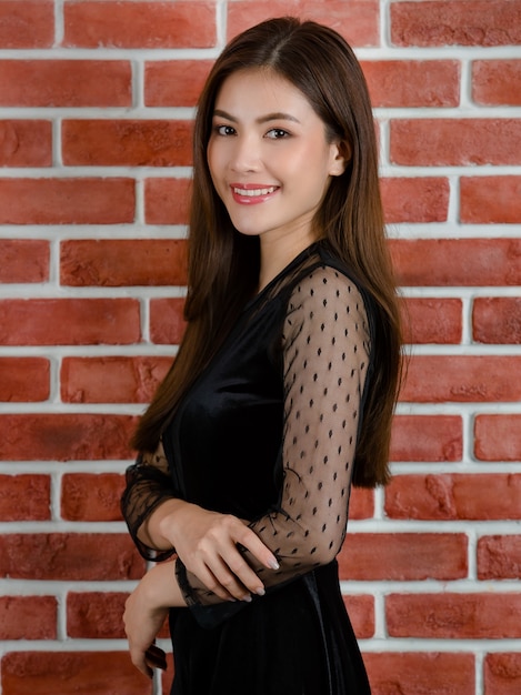 Portret młodej pięknej atrakcyjnej azjatyckiej modelki w czarnej sukni odsłaniając uśmiechając się i stojąc na tle pomarańczowego ceglanego muru.
