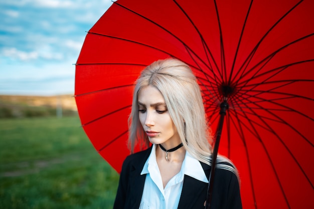 Portret młodej pięknej blondynki, trzymając czerwony parasol, ubrany w czarno-biały garnitur z obrożą na szyi. Zewnątrz tło niewyraźne zielone pole i zachmurzone niebo.