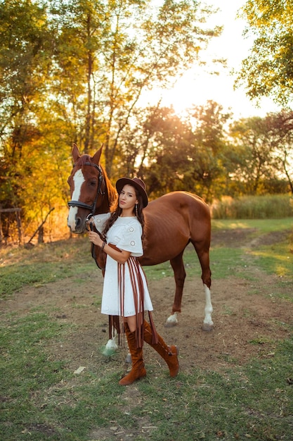 Portret młodej pięknej kobiety z kręconymi włosami, która trzyma brązowego konia Spacer ze zwierzętami na farmie Jazda konna Jeździec w stylu boho