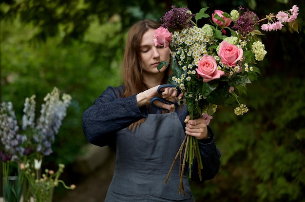 Portret młodej pięknej kwiaciarni w fartuchu na tarasie robi bukiet na zielonym tle