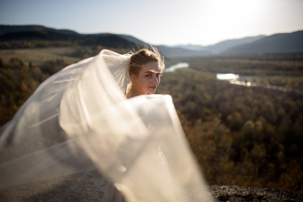 Portret młodej pięknej narzeczonej w górach z welonem. Wiatr rozwija zasłonę. Fotografia ślubna w górach.