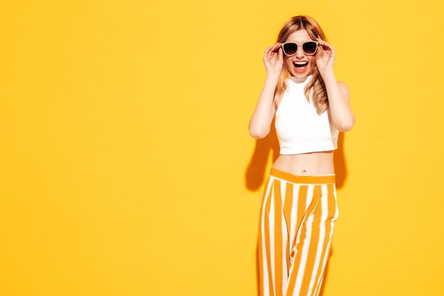 Portret młodej pięknej uśmiechniętej blond kobiety w modnych letnich ubraniach beztroska kobieta pozuje w pobliżu żółtej ściany w studio Pozytywny model zabawy w pomieszczeniu Wesoły i szczęśliwy w okularach przeciwsłonecznych