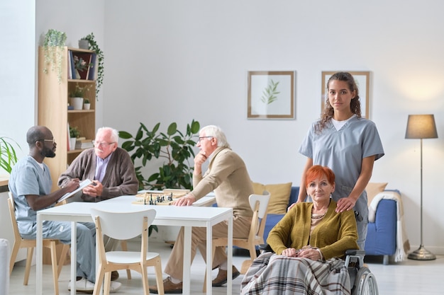 Portret młodej pielęgniarki ze starszą kobietą uśmiechającą się do kamery podczas pracy w domu opieki, która dba o seniorów