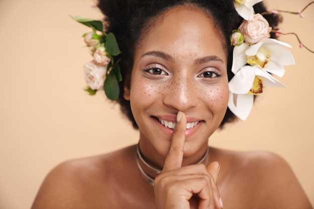 Portret młodej półnagiej kobiety z kwiatami we włosach robi cichy gest palcem na ustach