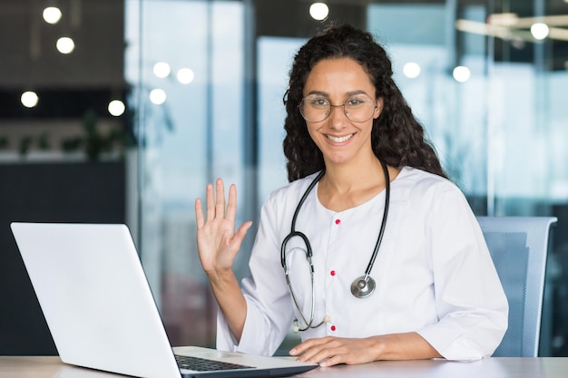 Portret młodej profesjonalnej lekarki z Ameryki Łacińskiej w białym fartuchu medycznym uśmiecha się i patrzy w kamerę trzymając rękę w górę gest powitania lekarka pracuje w środku