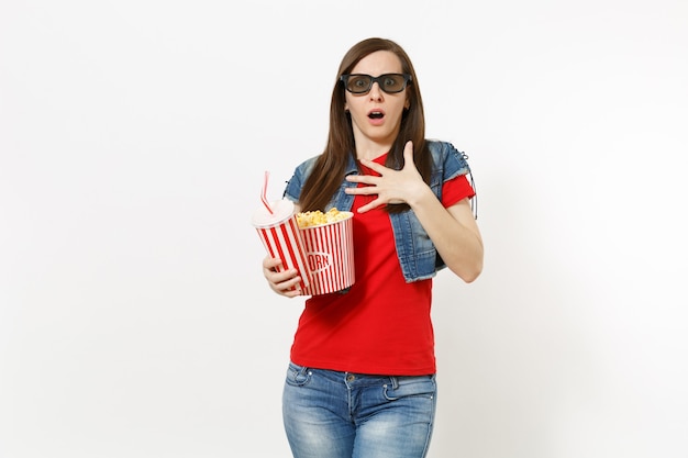Portret młodej przestraszonej kobiety w okularach 3d imax oglądania filmu, trzymając wiadro popcornu i plastikowy kubek napoju gazowanego lub coli, trzymając rękę na klatce piersiowej na białym tle. Emocje w kinie