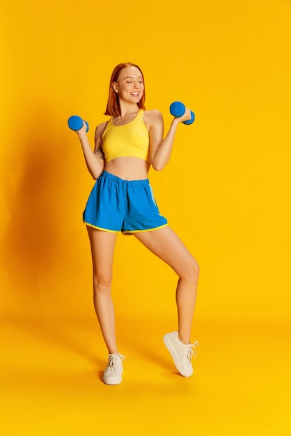 Portret młodej rudej dziewczyny w treningu odzieży sportowej ze sprzętem sportowym na żółtym tle Ćwiczenia rąk