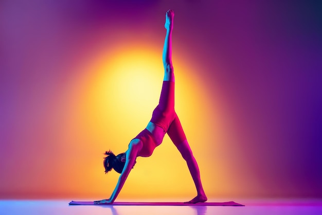 Portret młodej sportowej dziewczyny wykonującej ćwiczenia jogi podnoszącej nogę w górę, odizolowanej na różowym i żółtym tle gradientu w neonach