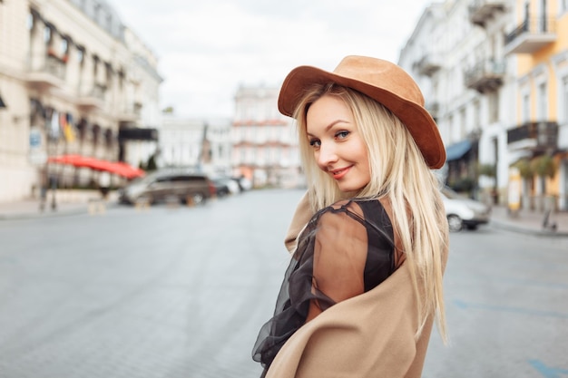 Portret młodej stylowej blondynki w jesiennym płaszczu i filcowym kapeluszu w europejskim mieście. Piękna atrakcyjna dziewczyna