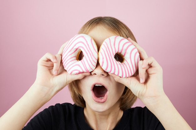 Zdjęcie portret młodej szczęśliwej blond dziewczyny trzymającej dwa pączki na oczach i patrząc przez nie. pojęcie niezdrowego uzależnienia od słodkiego odżywiania.