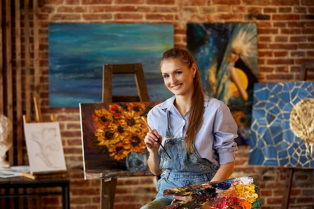 Portret młodej uśmiechniętej artystki z jej dziełami sztuki w studiu artystycznym malarza trzymającego pędzle artystyczne