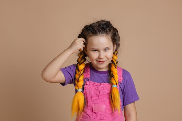 Portret młodej uśmiechniętej dziewczyny z warkoczami kanekalon żółtego koloru dotykającymi włosów ręką patrząc na kamerę w różowym kombinezonie i fioletowej koszulce na beżowym tle