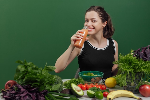Portret młodej wesołej kobiety ze świeżym sokiem przy stole pełnym zdrowych surowych warzyw i ...