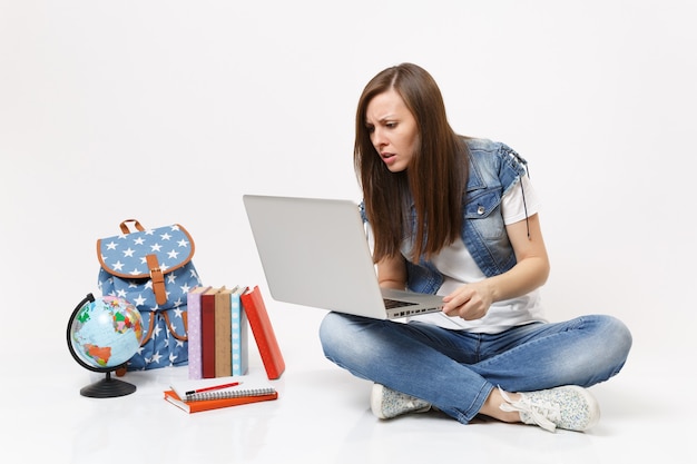 Portret młodej zaniepokojonej, zaintrygowanej studentki trzymającej się za pomocą laptopa, siedzącej w pobliżu kuli ziemskiej, plecaka, podręczników szkolnych na białym tle