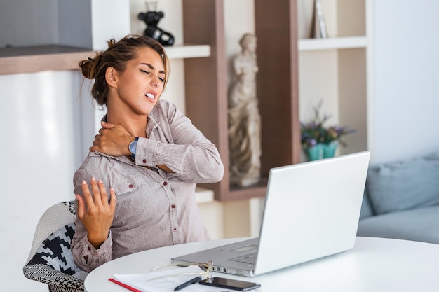 Portret młodej zestresowanej kobiety siedzącej przy biurku w domu przed laptopem, dotykającej bólu pleców z bolesnym wyrazem twarzy, cierpiącej na ból pleców po pracy na komputerze