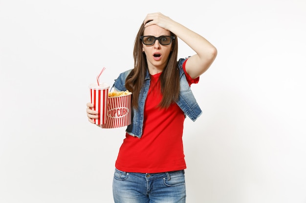 Portret młodej zszokowanej kobiety w okularach 3d oglądając film, trzymając wiadro popcornu i plastikowy kubek napoju gazowanego lub coli, trzymając się głowy na białym tle. Emocje w koncepcji kina.
