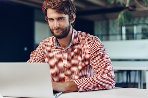 Portret młody caucasian biznesmen używa laptop przy jego miejscem pracy w nowożytnym biurze