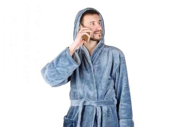 Portret młody caucasian brodaty mężczyzna w błękitnym bathrobe opowiada na smartphone odizolowywającym na białym tle