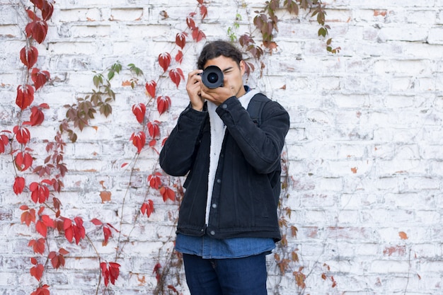 Portret młody początkujący fotograf stoi blisko biel ściany