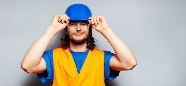 Portret młody ufny pracownik budowlany jest ubranym sprzęt ochronny; niebieski kask, przezroczyste okulary i żółta kamizelka.