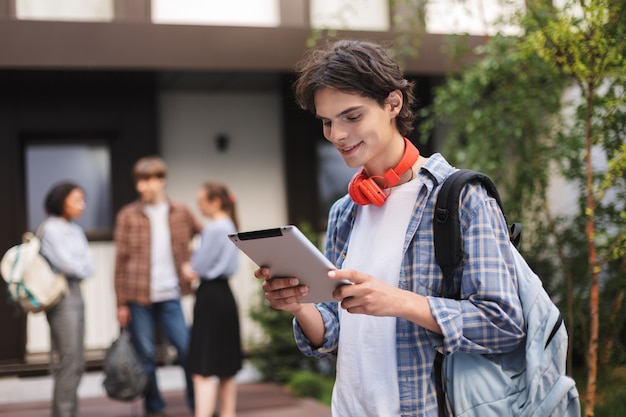 Portret Młodzieńca Z Czerwonymi Słuchawkami I Plecakiem, Stojąc I Za Pomocą Tabletu, Spędzając Czas Na Dziedzińcu Uniwersytetu Ze Studentami Na Tle