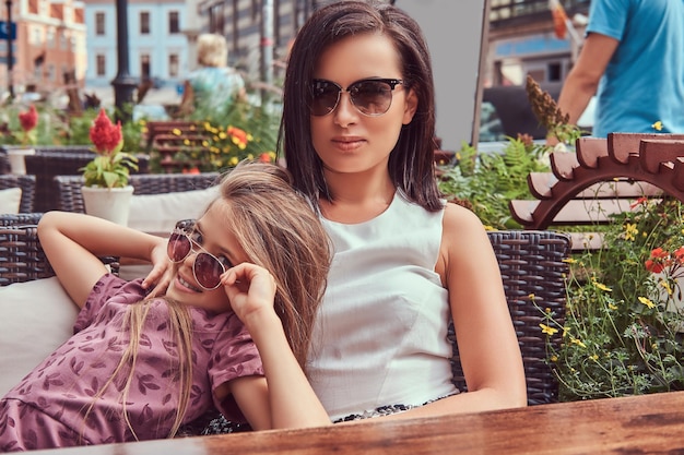 Portret modnej mamy i jej uroczej córki w okularach przeciwsłonecznych w kawiarni na świeżym powietrzu. Szczęśliwa modna rodzina.