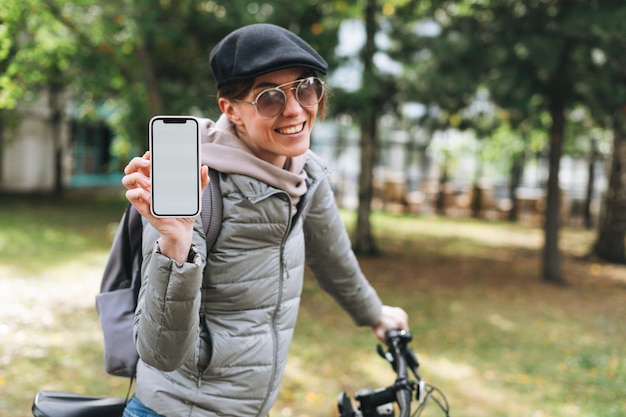 Portret modnej szczęśliwej młodej ładnej kobiety w czapce i okularach przeciwsłonecznych za pomocą telefonu komórkowego na rowerze w słoneczny jesienny dzień w parku miejskim Makieta mobilna smartphone z białym ekranem