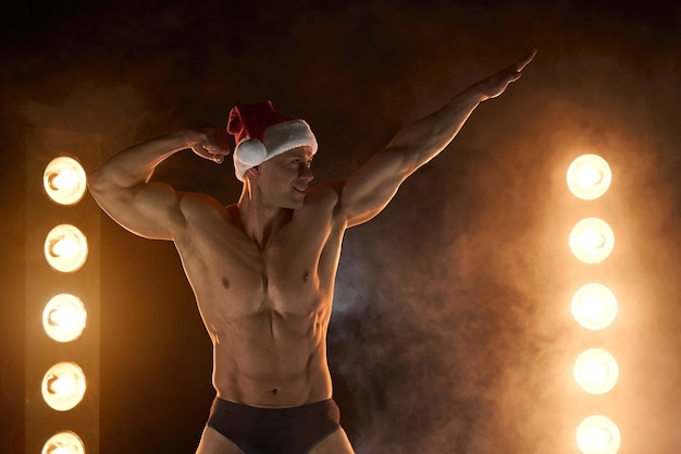 Portret muskularnego mężczyzny w świątecznej czapce Mikołaja, stojącej atletycznej pozie na zadymionym tle Macho Shirtless Naga striptizerka torsu Hot sexy Santa guy Uwodzicielski mężczyzna na wakacje